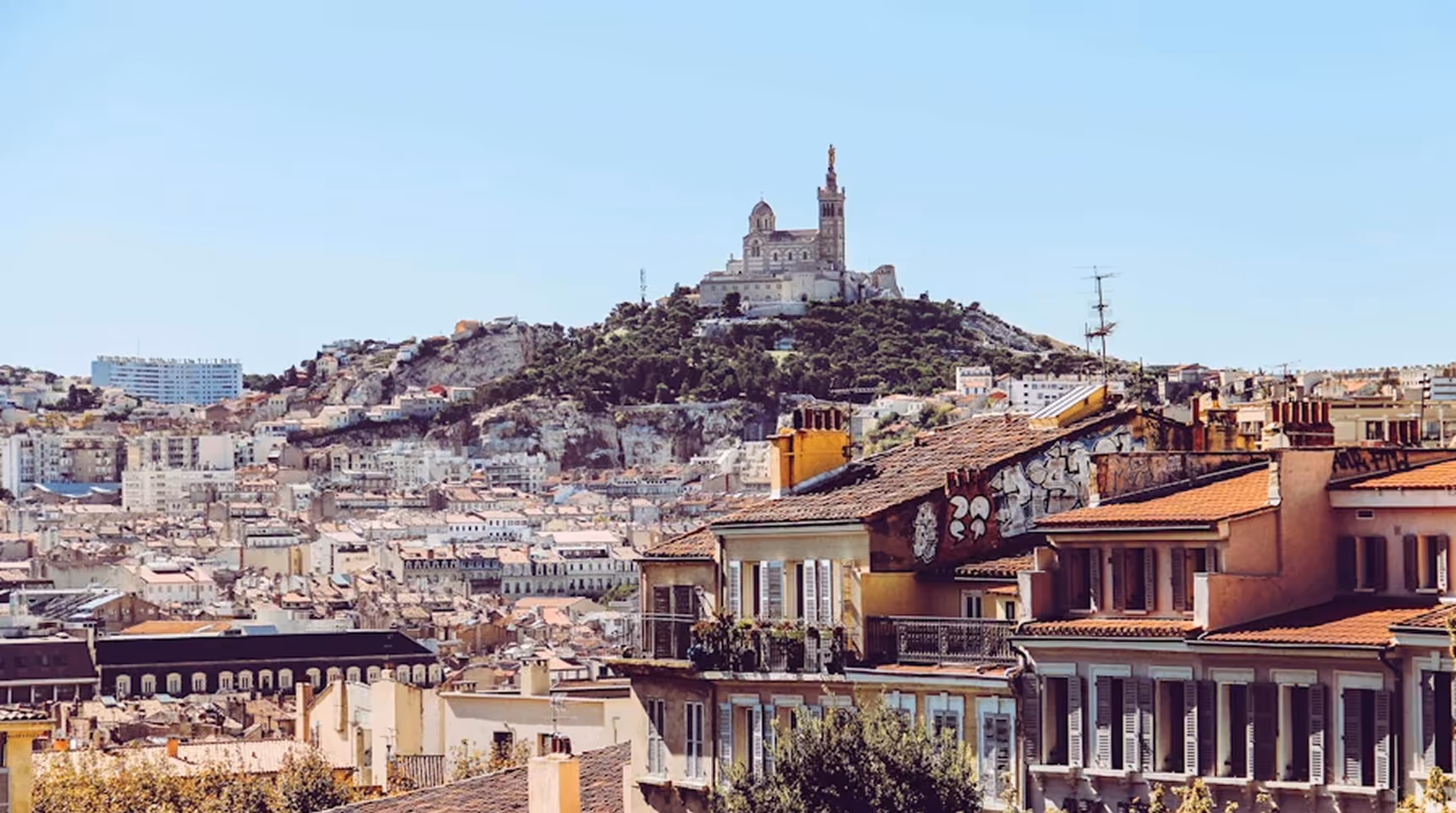 Cityscape of Marseille with terracotta rooftops and Notre-Dame de la Garde basilica atop a hill under a clear blue sky.
