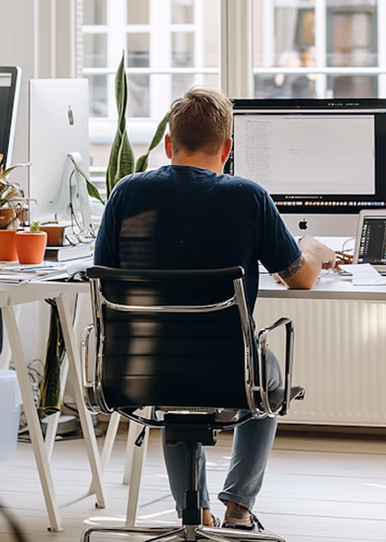 Person sitting at a desk working on an iMac computer in a bright office with plants and papers around.