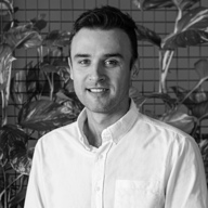 Smiling man with short dark hair wearing a white button-up shirt, standing in front of a grid-patterned wall with leafy plants.