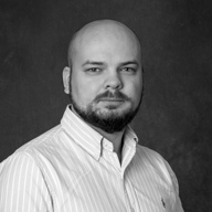 Portrait of a bald man with a beard wearing a striped button-up shirt against a dark background.