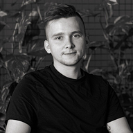Black and white portrait of a young man with short hair wearing a black t-shirt, sitting in front of a brick wall with plants.