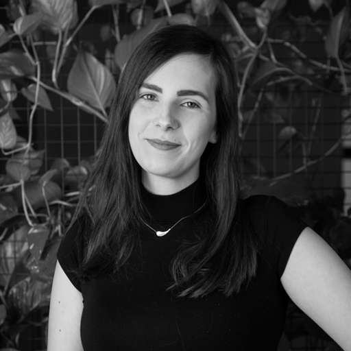Portrait of a smiling woman with long dark hair wearing a black top and necklace standing in front of leafy plants.