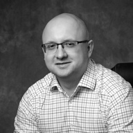 Portrait of a bald man wearing glasses and a checkered shirt, smiling against a plain dark background.