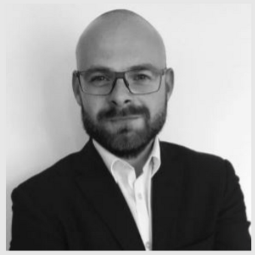 Portrait of a bald man with glasses and a beard wearing a dark suit and white shirt.