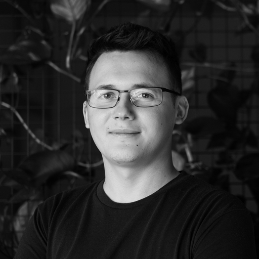 Portrait of a young man wearing glasses and a black t-shirt, with a plant-filled background.