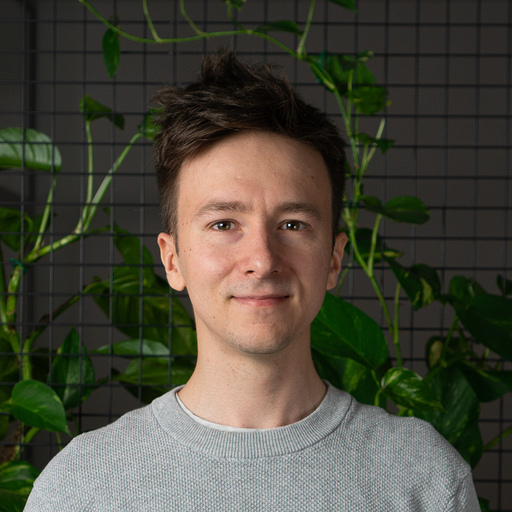 Young man with short dark hair wearing a gray sweater, standing in front of a black grid wall with green plants.