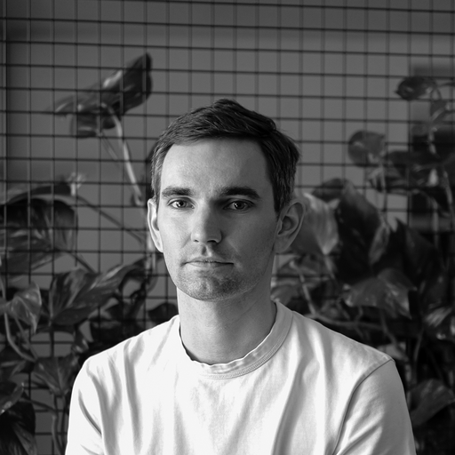 Black and white portrait of a man with short dark hair wearing a plain white shirt, standing in front of a grid wall with plants.