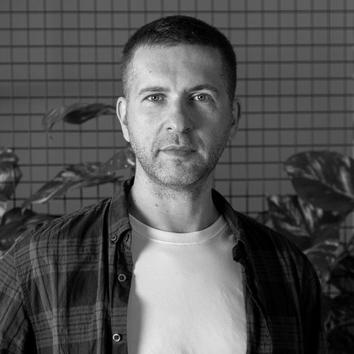 Black and white portrait of a man with short hair wearing a plaid shirt over a white t-shirt, standing in front of a grid wall with plants.