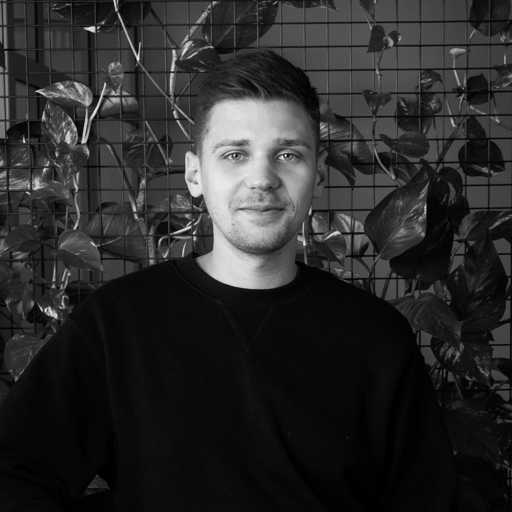 Black and white portrait of a young man with short hair wearing a black sweater, standing in front of a grid wall with leafy plants.