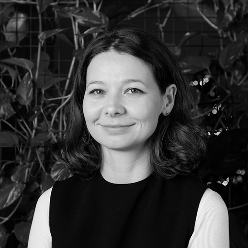 Smiling woman with shoulder-length hair wearing a sleeveless top over a long-sleeve shirt standing in front of leafy plants.