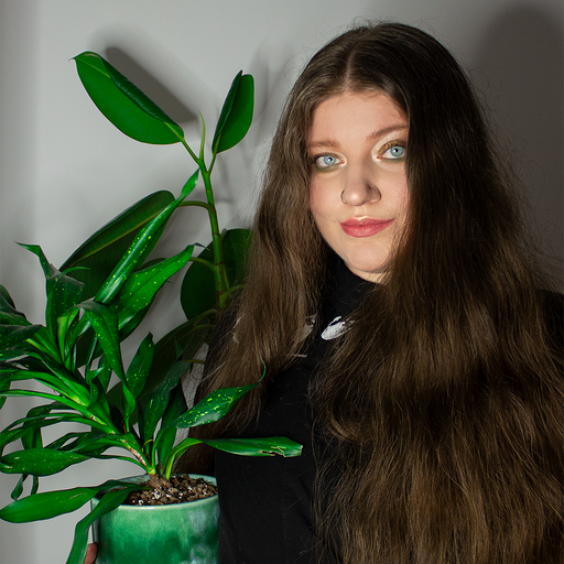 Young woman with long brown hair and blue eyes wearing a black shirt, holding a green potted plant with broad leaves.
