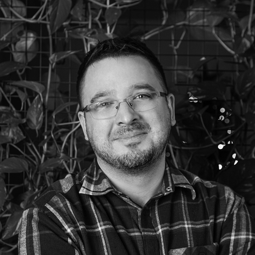 Smiling man with glasses and a beard wearing a plaid shirt standing in front of a leafy background.
