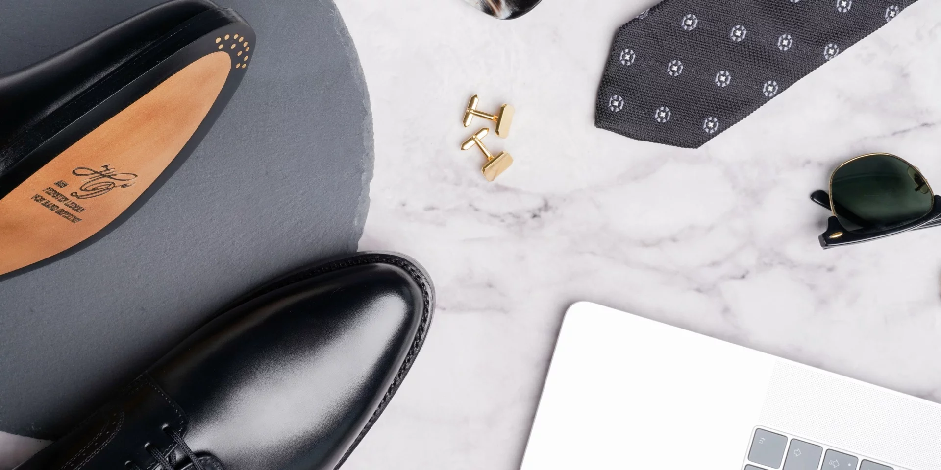 Flat lay of men's black leather dress shoes, gold cufflinks, patterned necktie, sunglasses, and a laptop on a marble surface.
