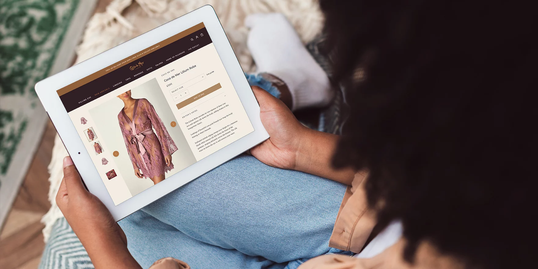 Woman holding a tablet showing an online shopping page for a pink lace Coco de Mer Lilium Robe.