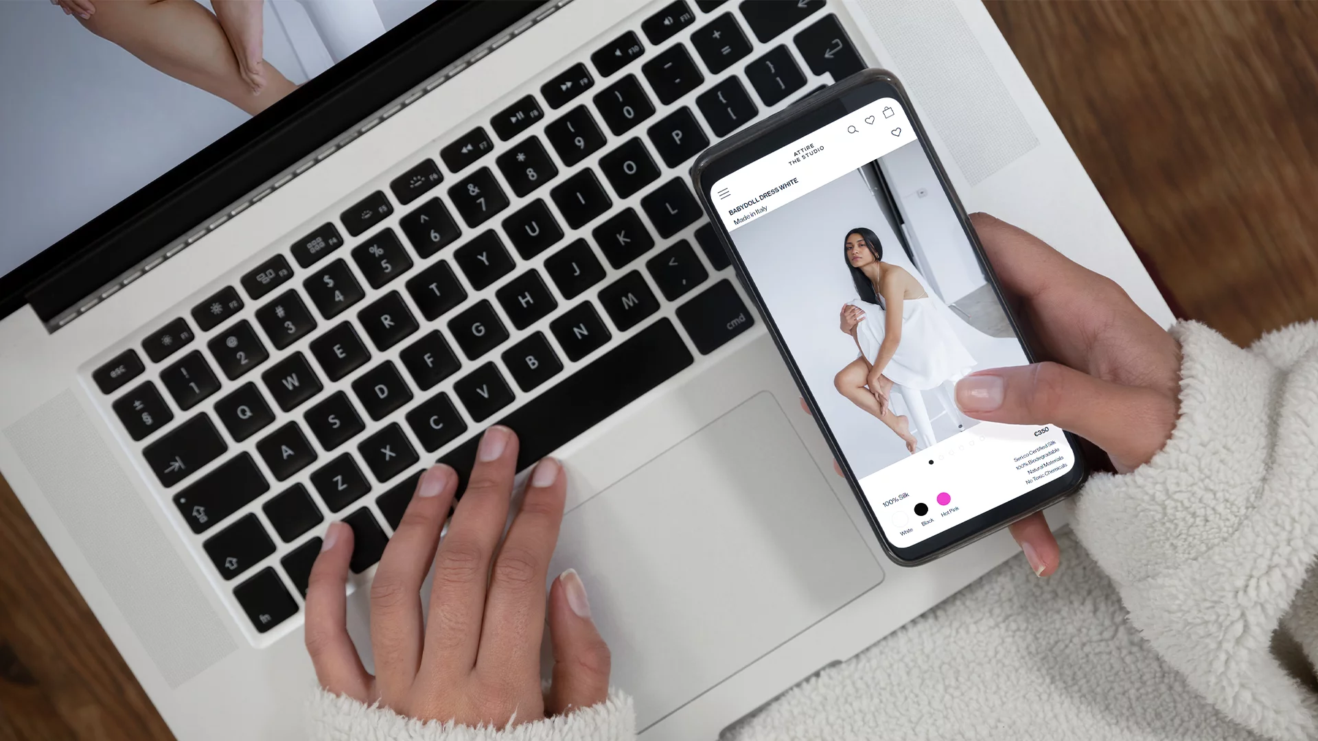 Person using a laptop with one hand on the trackpad and holding a smartphone displaying an online fashion store featuring a woman in a white dress.