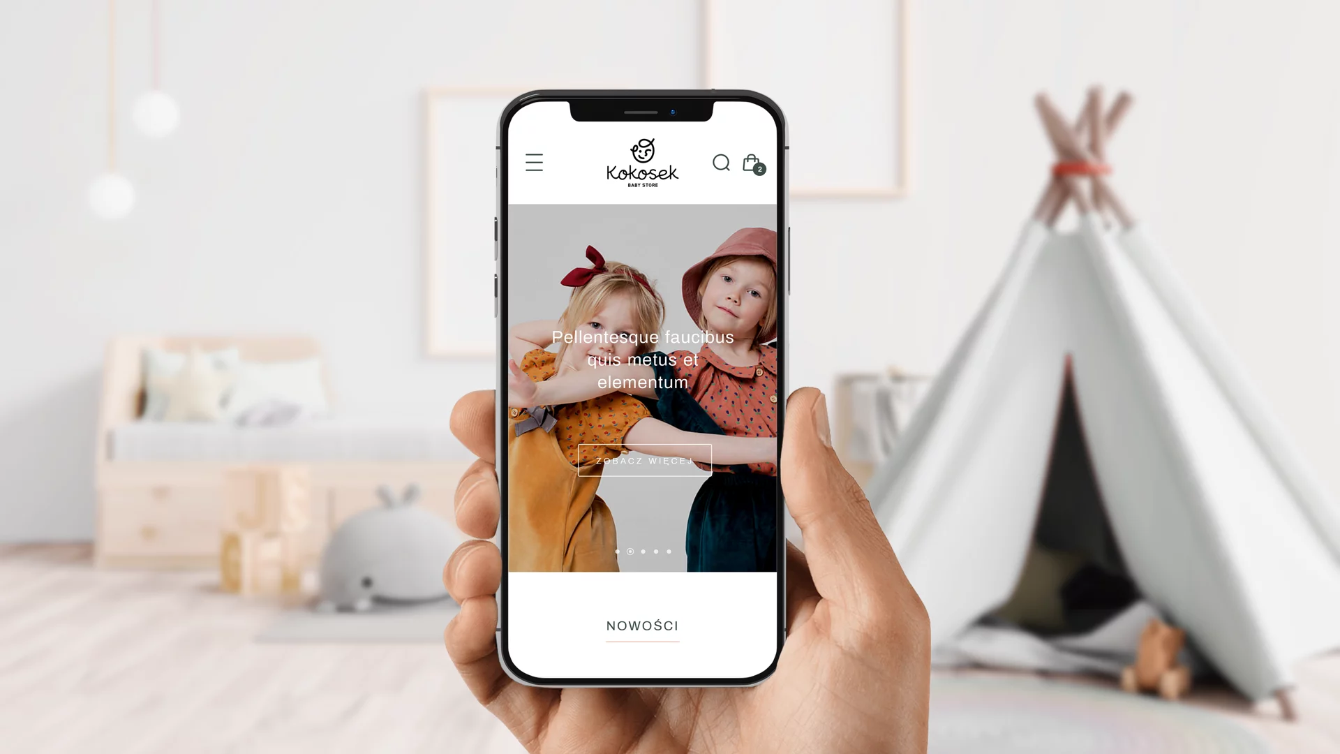 Hand holding a smartphone displaying Kokosek baby store website with two children posing on screen, blurred nursery background with bed and teepee.