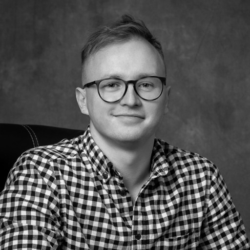 Young man with short hair and glasses wearing a checkered shirt, smiling slightly while sitting in a chair.