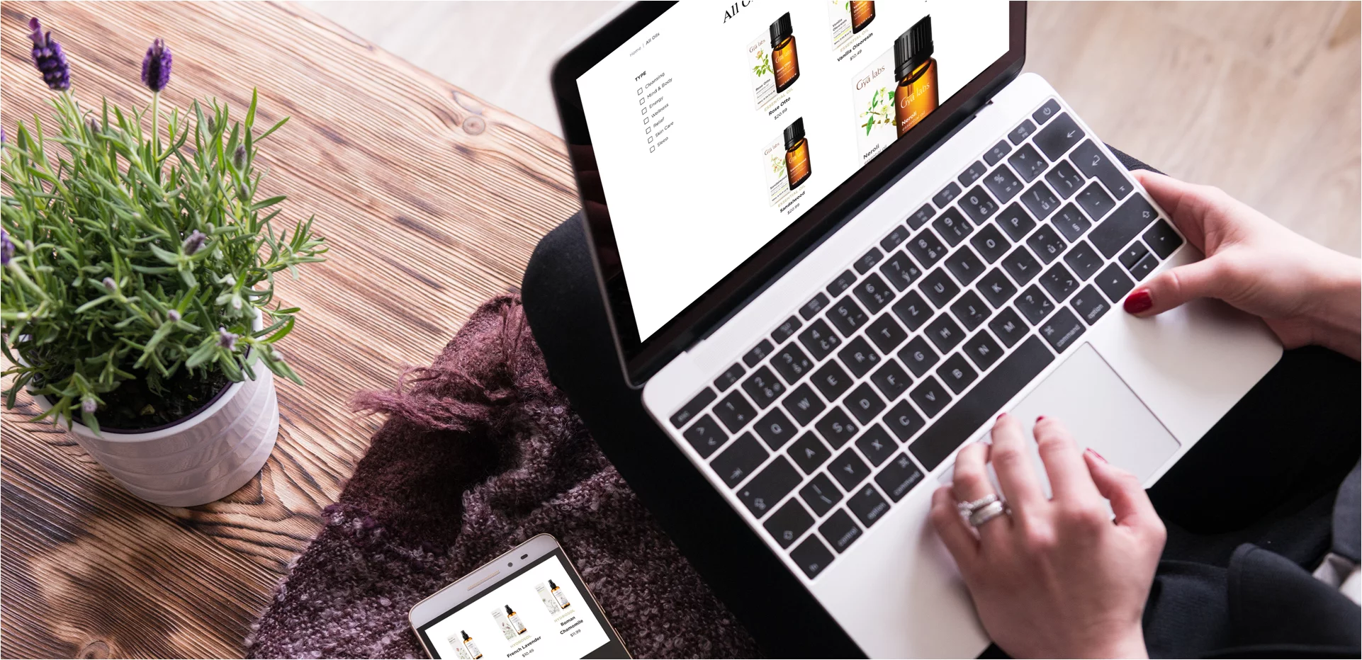Hands with red nail polish using a laptop displaying essential oil products, next to a smartphone with similar product images and a potted lavender plant on a wooden surface.