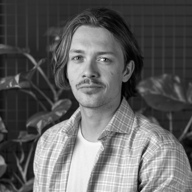 Black and white portrait of a young man with shoulder-length hair and a mustache, wearing a plaid shirt over a white t-shirt, against a background with plants and a grid wall.