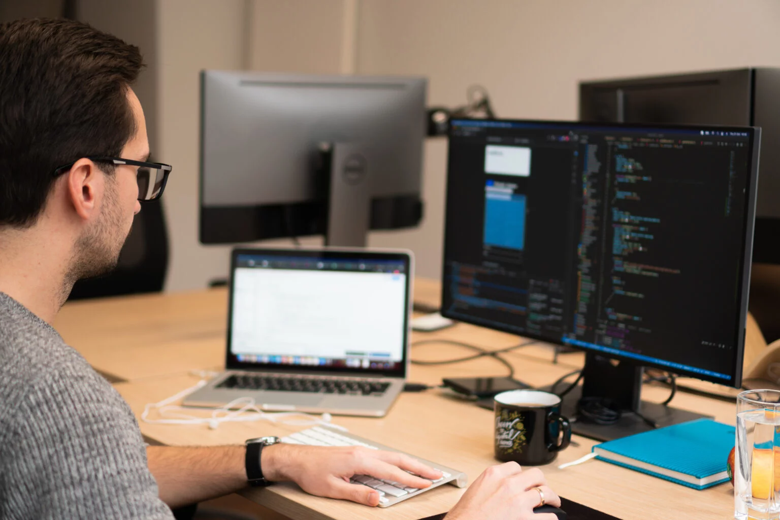 Man wearing glasses working on a laptop and large monitor displaying code at a desk with a keyboard, coffee mug, notebook, and glass of water.