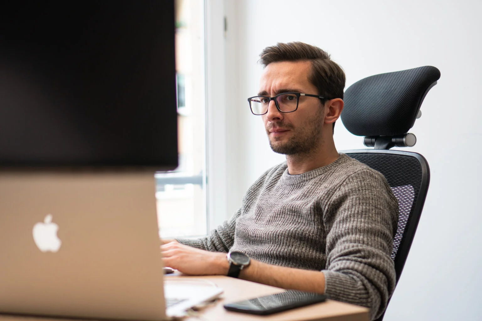 Man with glasses and a gray sweater sitting in an office chair, focused on a large Apple computer screen.
