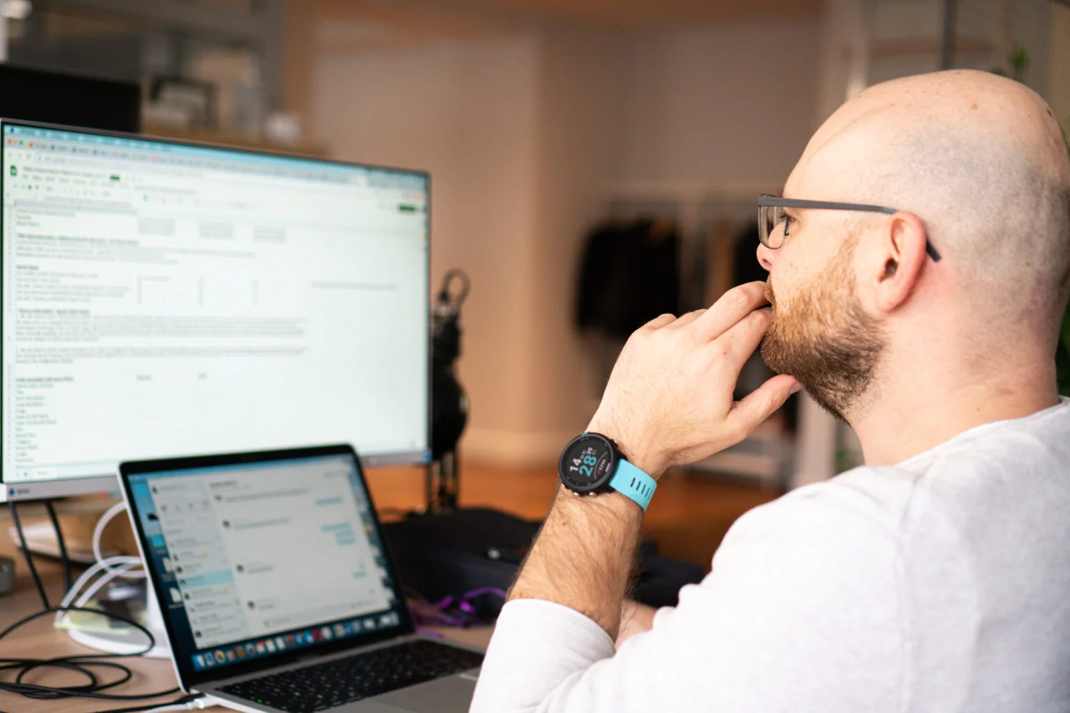 Bald man with glasses and beard wearing a blue smartwatch, thoughtfully looking at two computer screens on a desk.