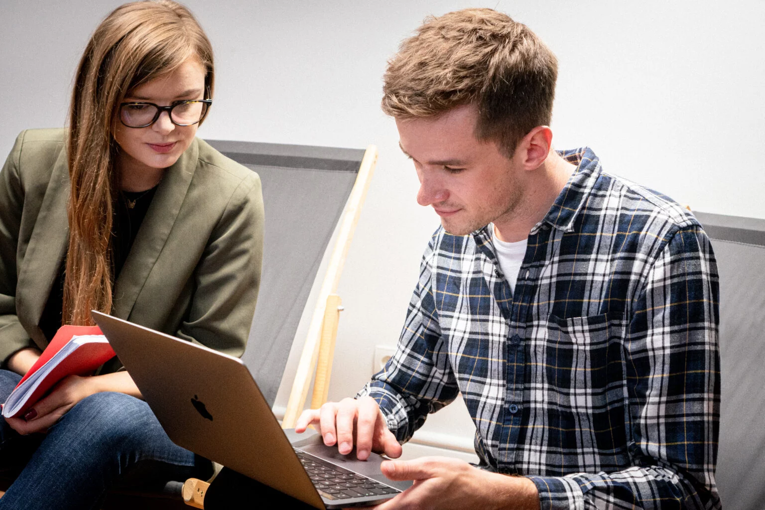 A man in a checkered shirt working on a laptop while a woman with glasses and a green blazer watches, holding a red notebook.