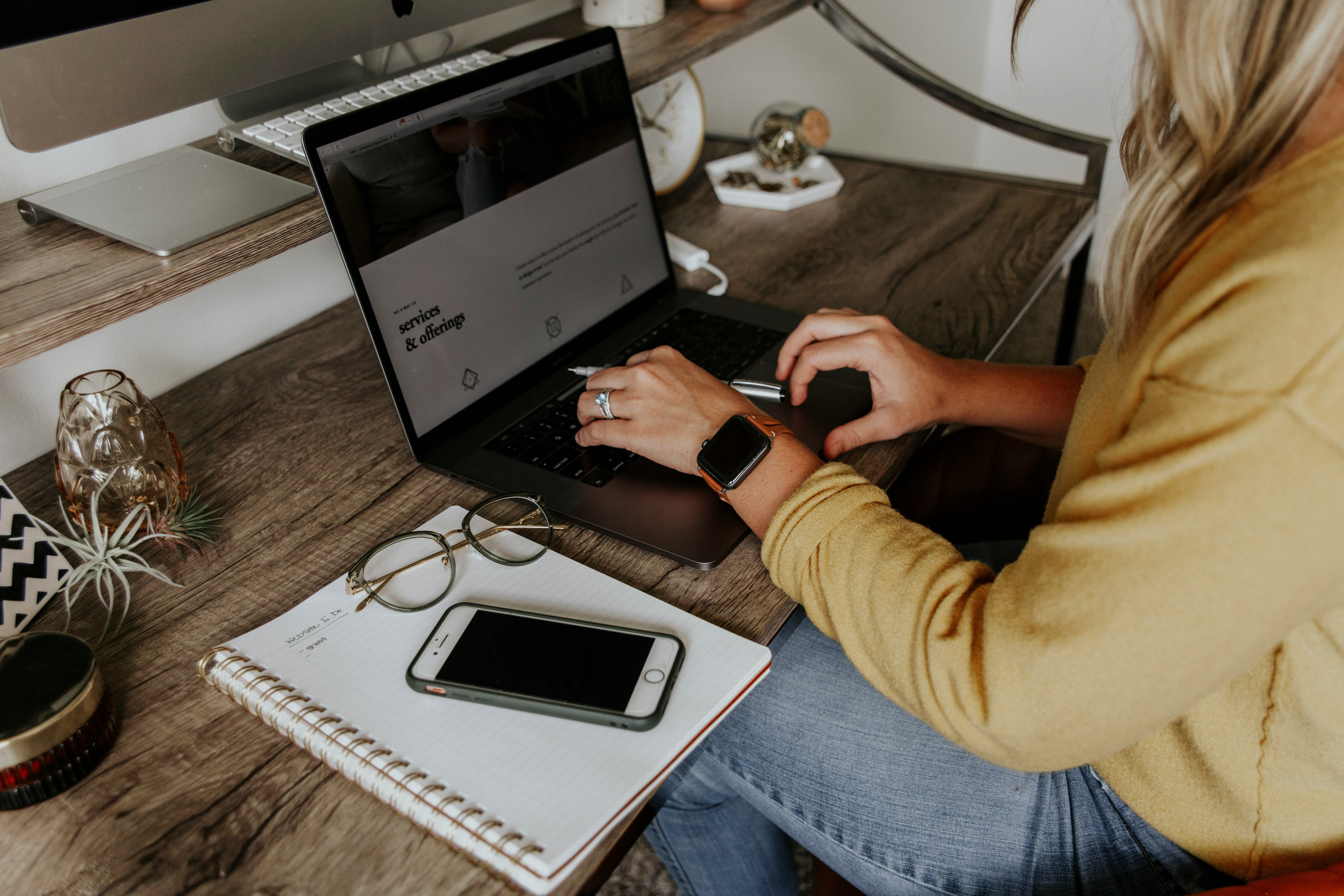 Woman typing on a laptop at a wooden desk with a phone, glasses and notebook, preparing a store for AI-driven sales