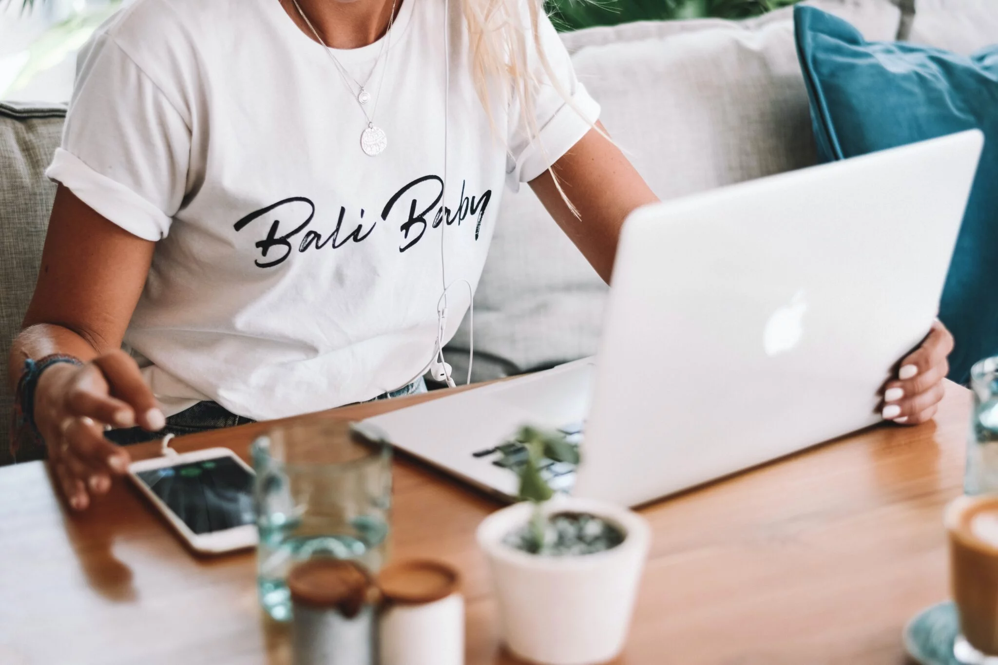 Woman in a white 'Bali Baby' T-shirt working on a MacBook at a café table with a phone and succulent