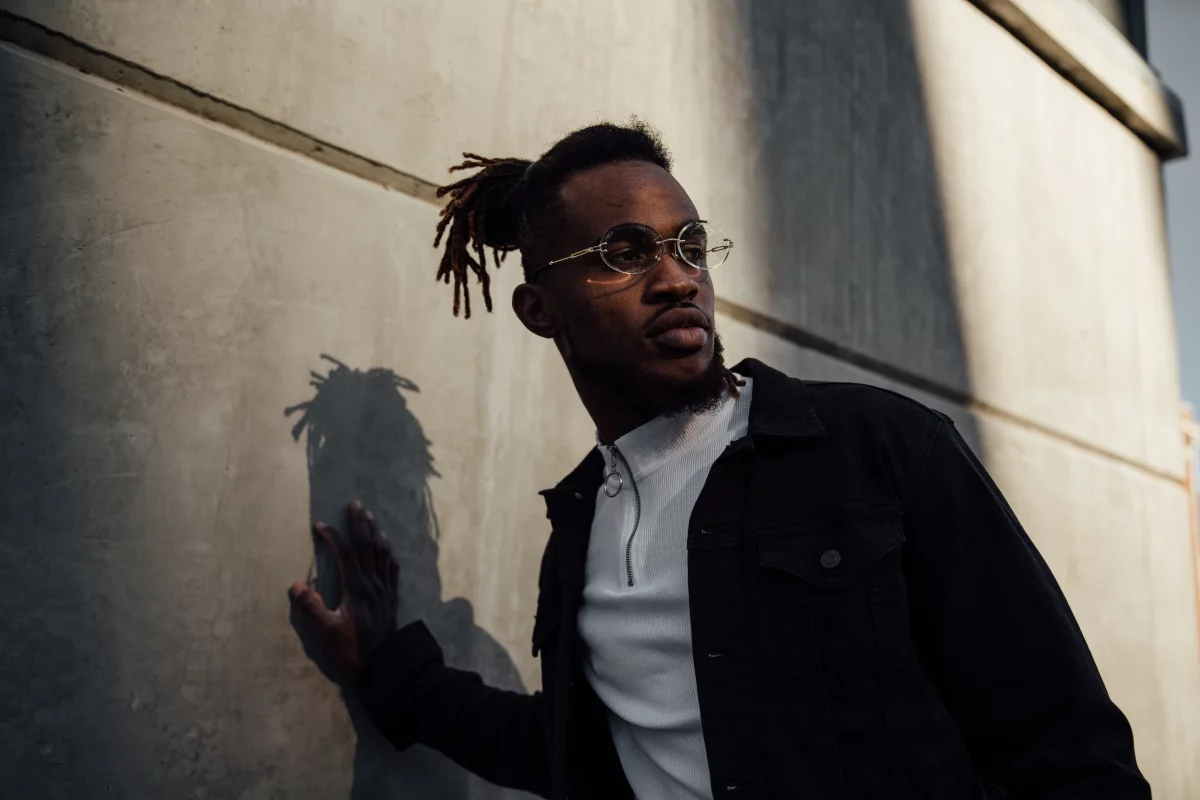 Stylish man in round glasses and a black denim jacket leaning on a concrete wall, evoking a fashion brand on Shopify