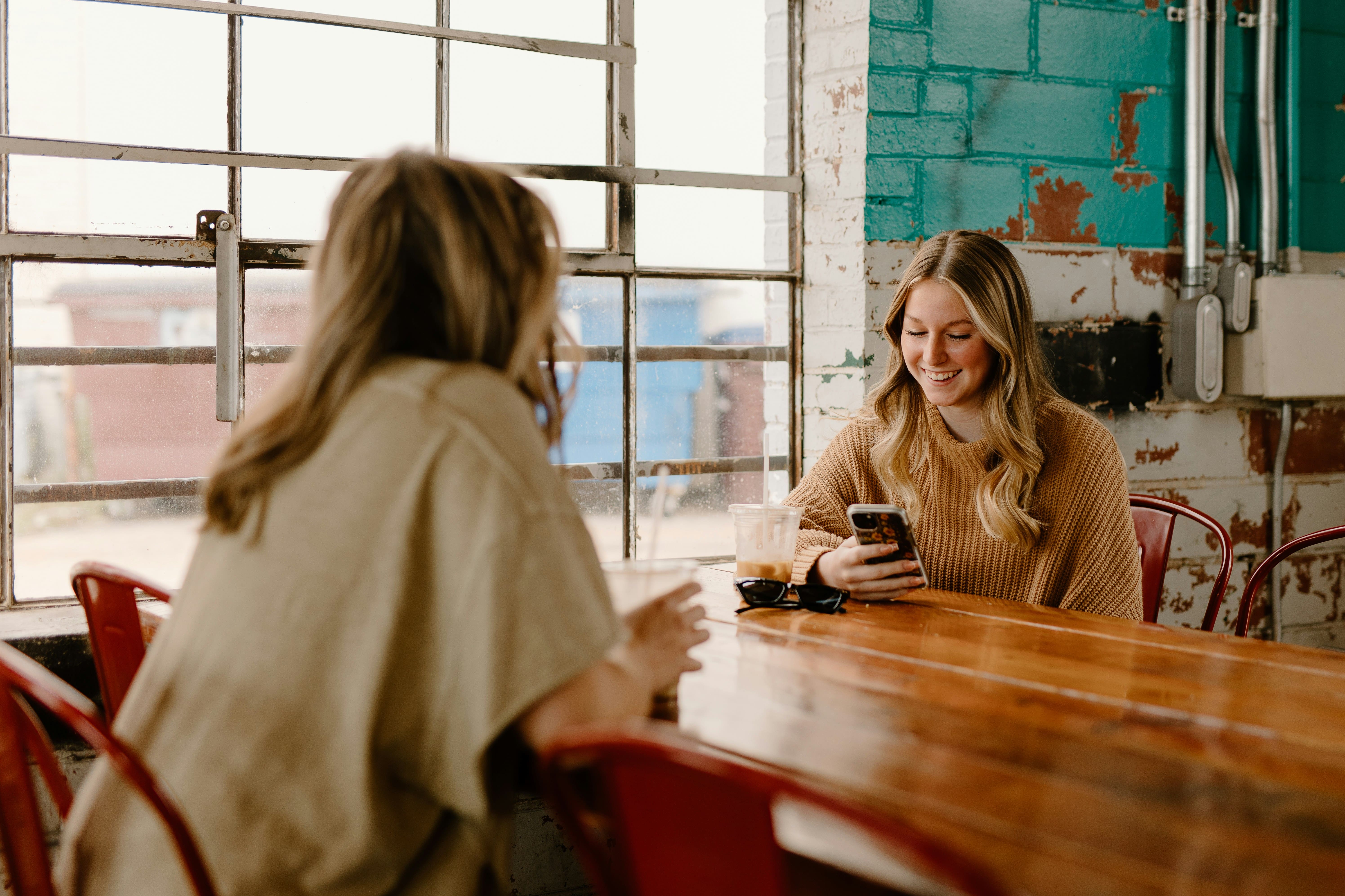 Two women at a wooden café table, one smiling at her phone, illustrating mobile shopping for a fashion brand