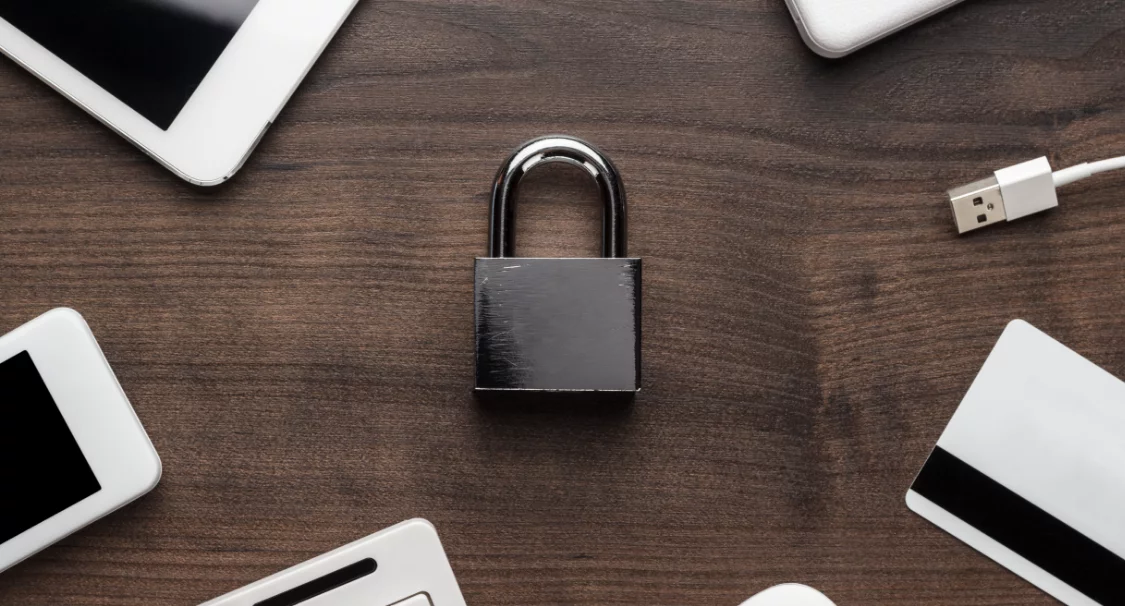 Black padlock on a wooden desk surrounded by a tablet, phone, USB cable and payment card, symbolising store security