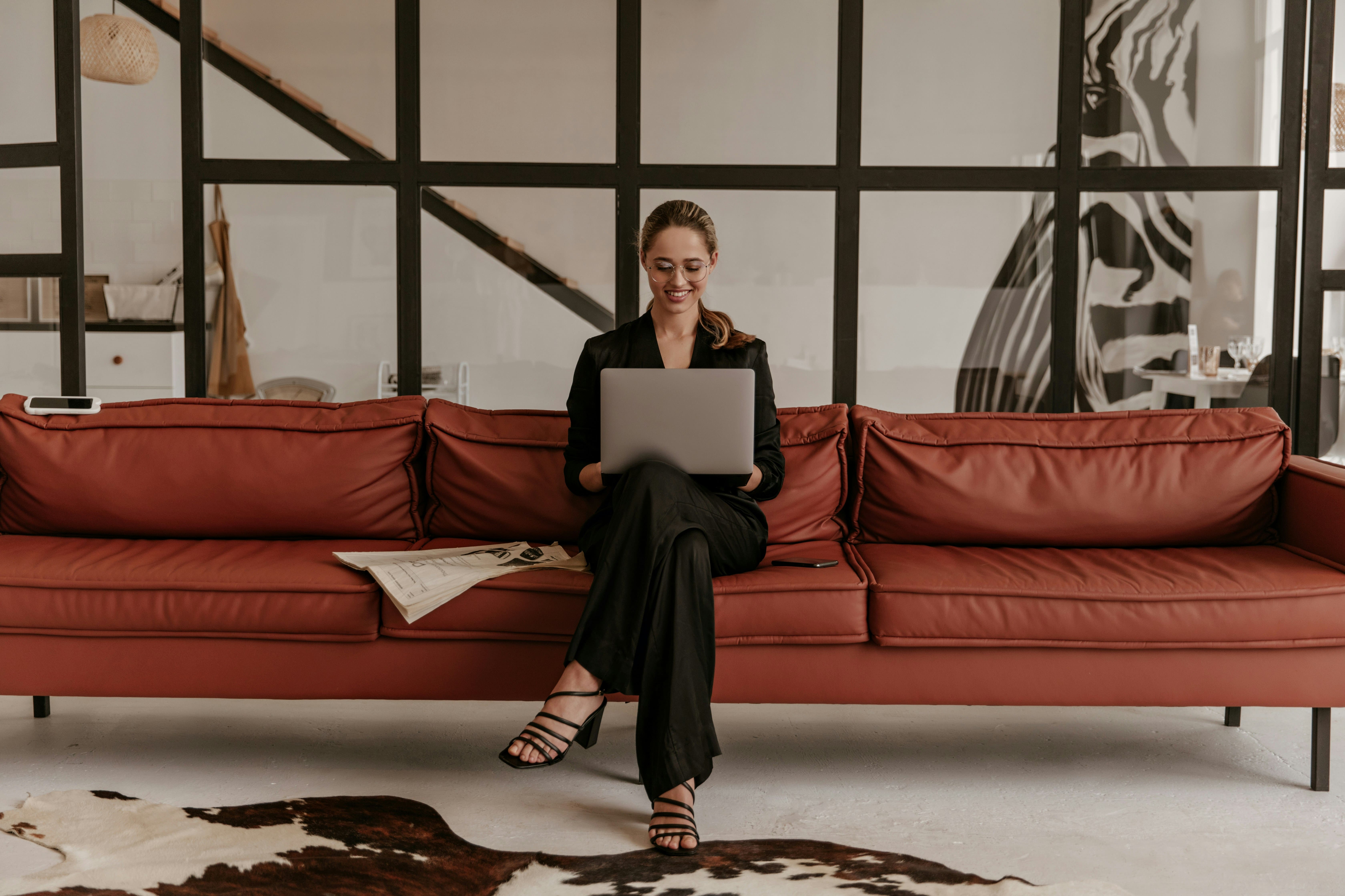 Woman in black sitting on a rust-red leather sofa with a laptop in a stylish interior, evoking a furniture brand