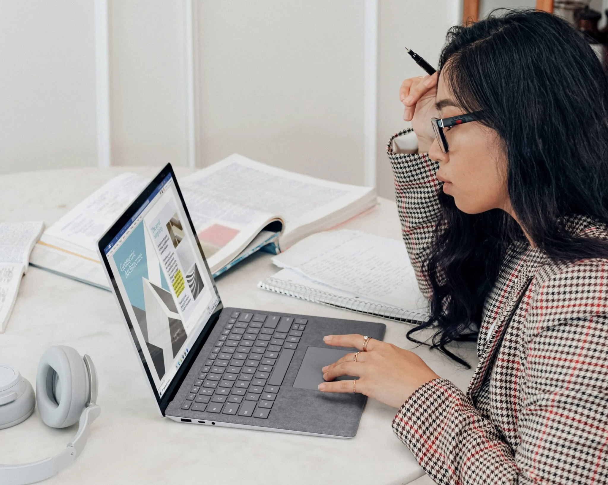Woman in glasses and a houndstooth blazer working on a laptop among open books, researching fashion brand strategy