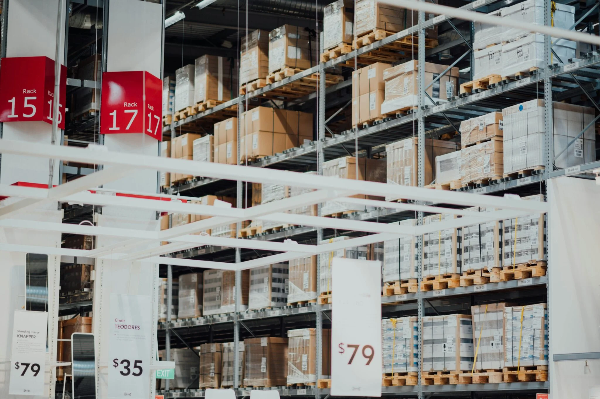 Furniture warehouse with tall racks of boxed goods on pallets, numbered rack signs and price tags
