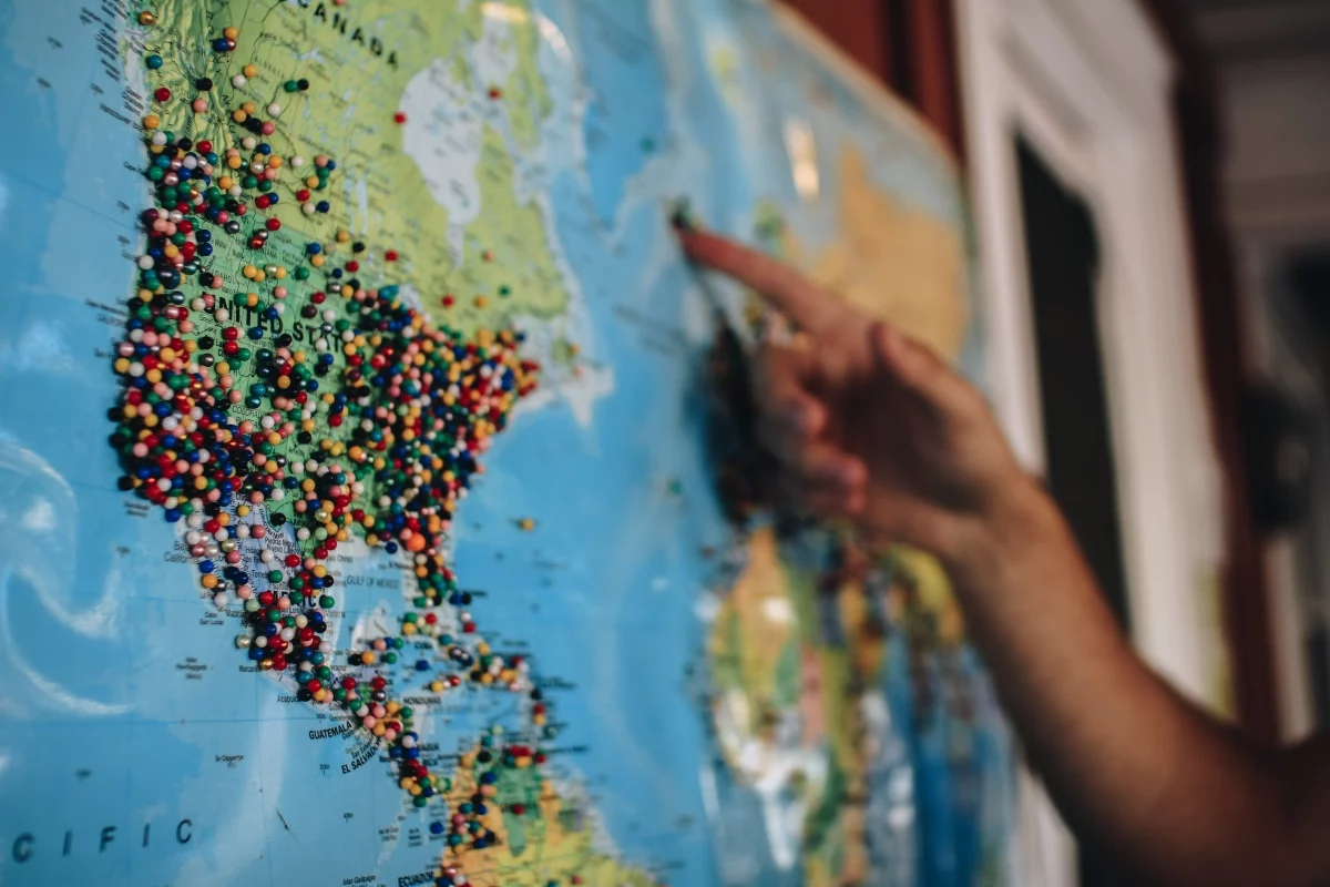 Hand pointing at a world map covered with colourful pins across North America, illustrating selling abroad
