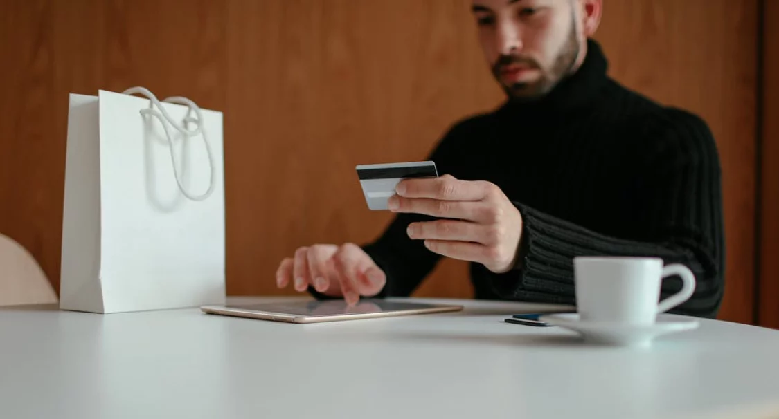Man holding a payment card while tapping a tablet next to a white shopping bag and coffee cup, paying online