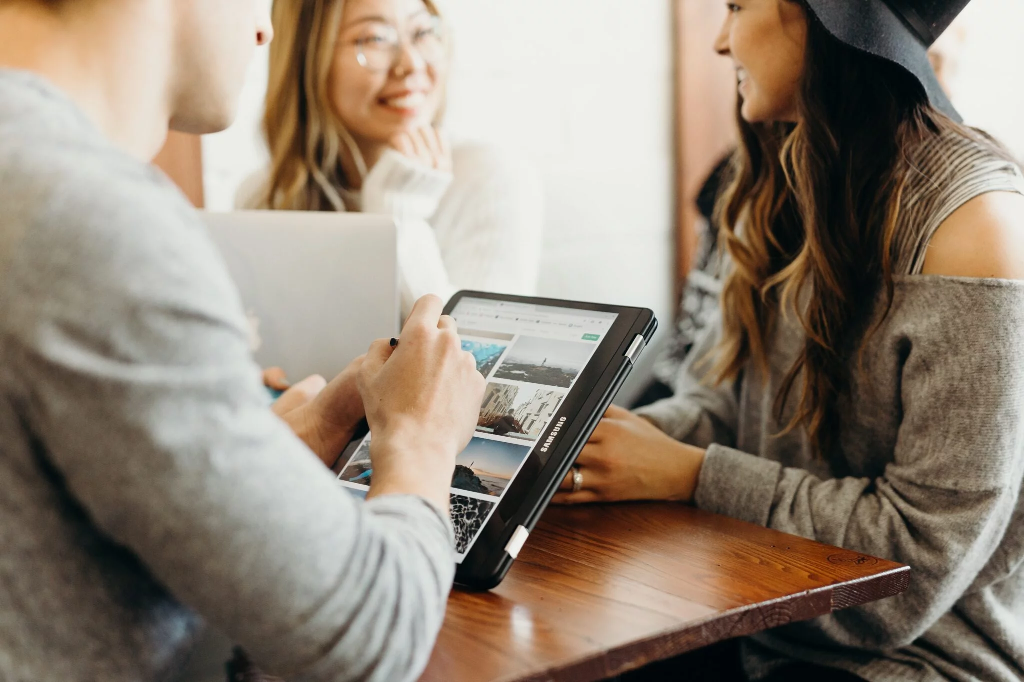 Three friends at a café table smiling while one browses photos on a tablet, evoking loyal customer relationships