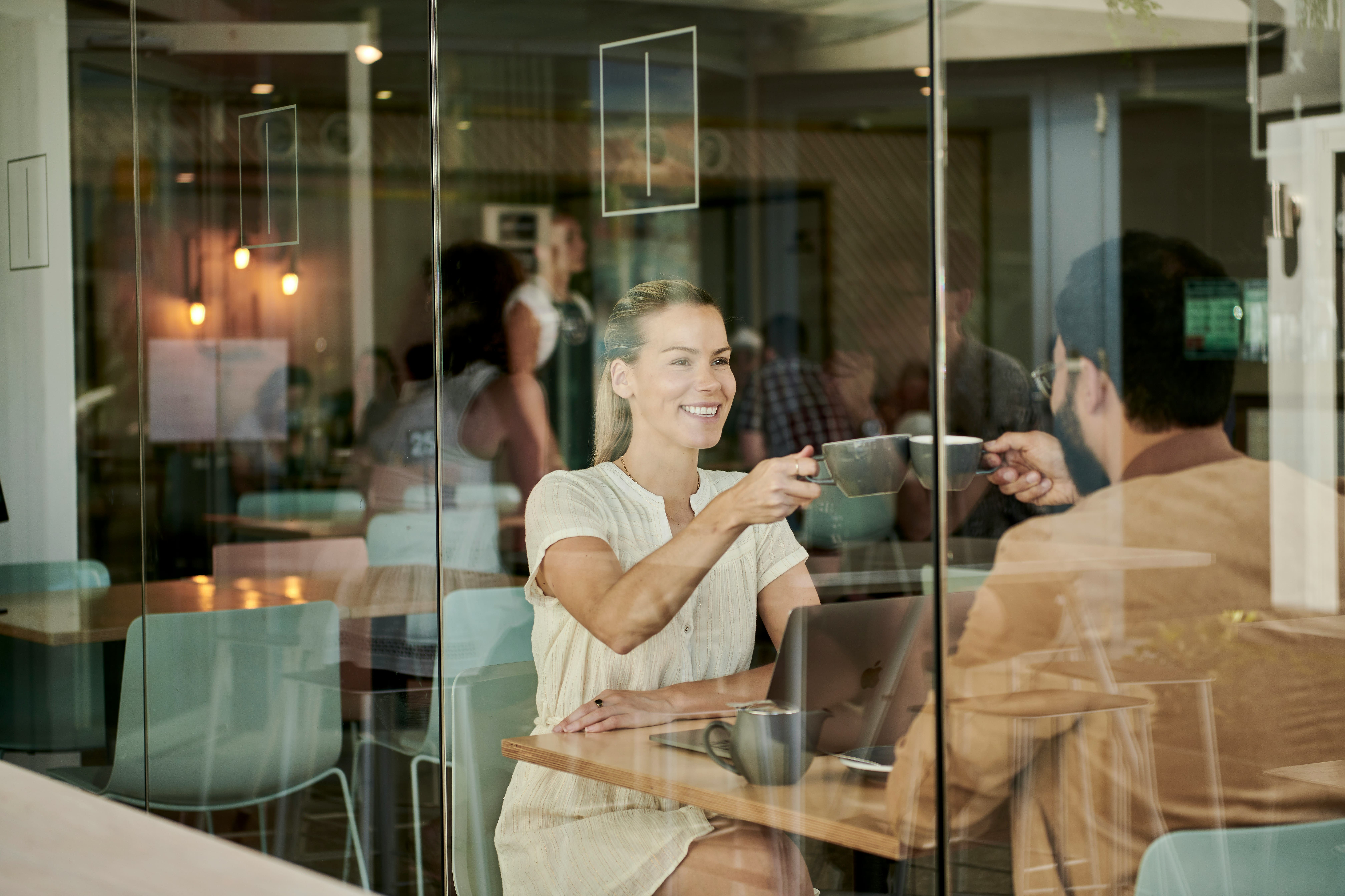 Woman and man toasting with coffee cups at a laptop in a glass-walled café, illustrating returning customers