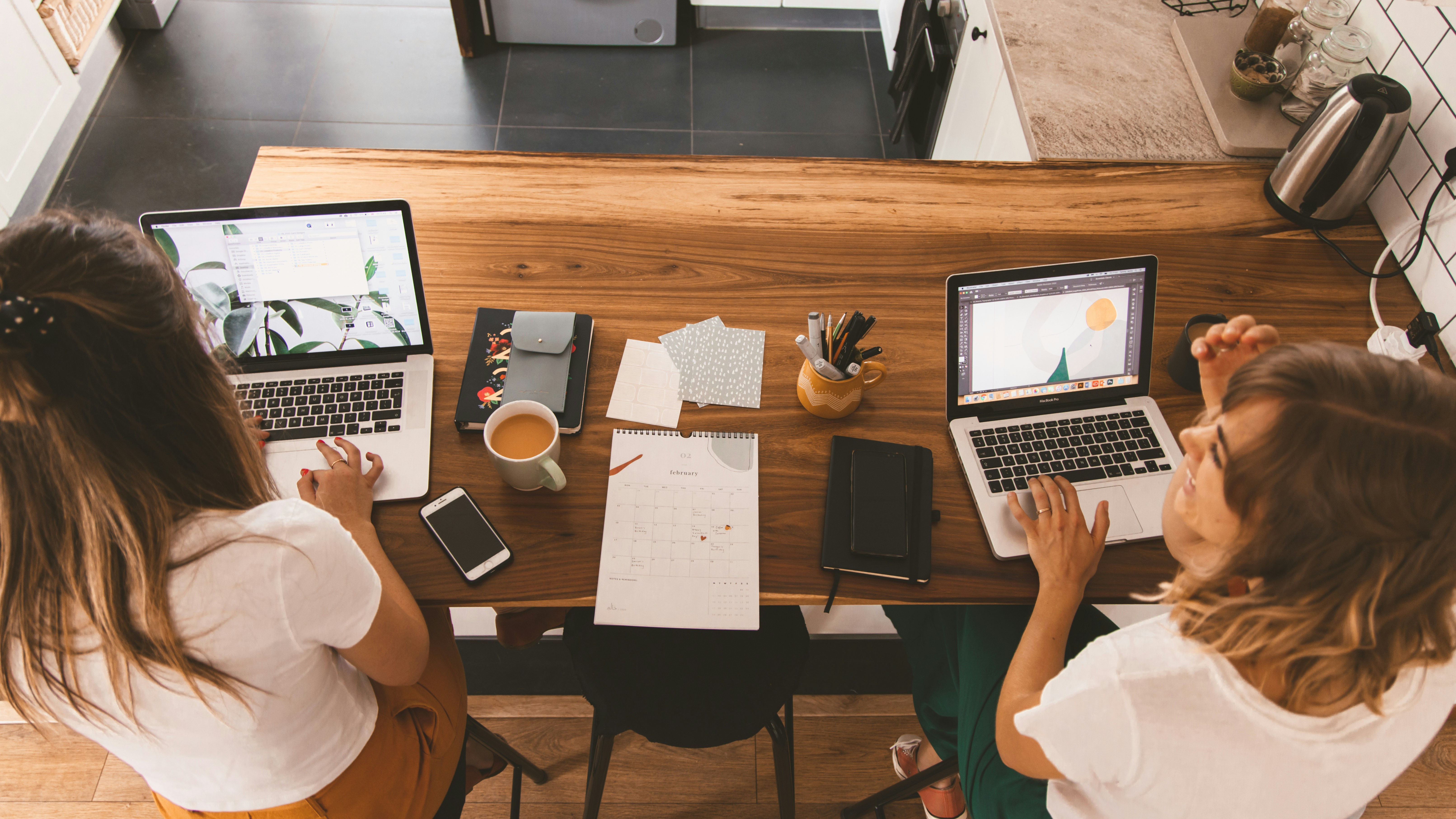 Two women working on laptops at a wooden kitchen table with notebooks and tea, reviewing data about existing customers