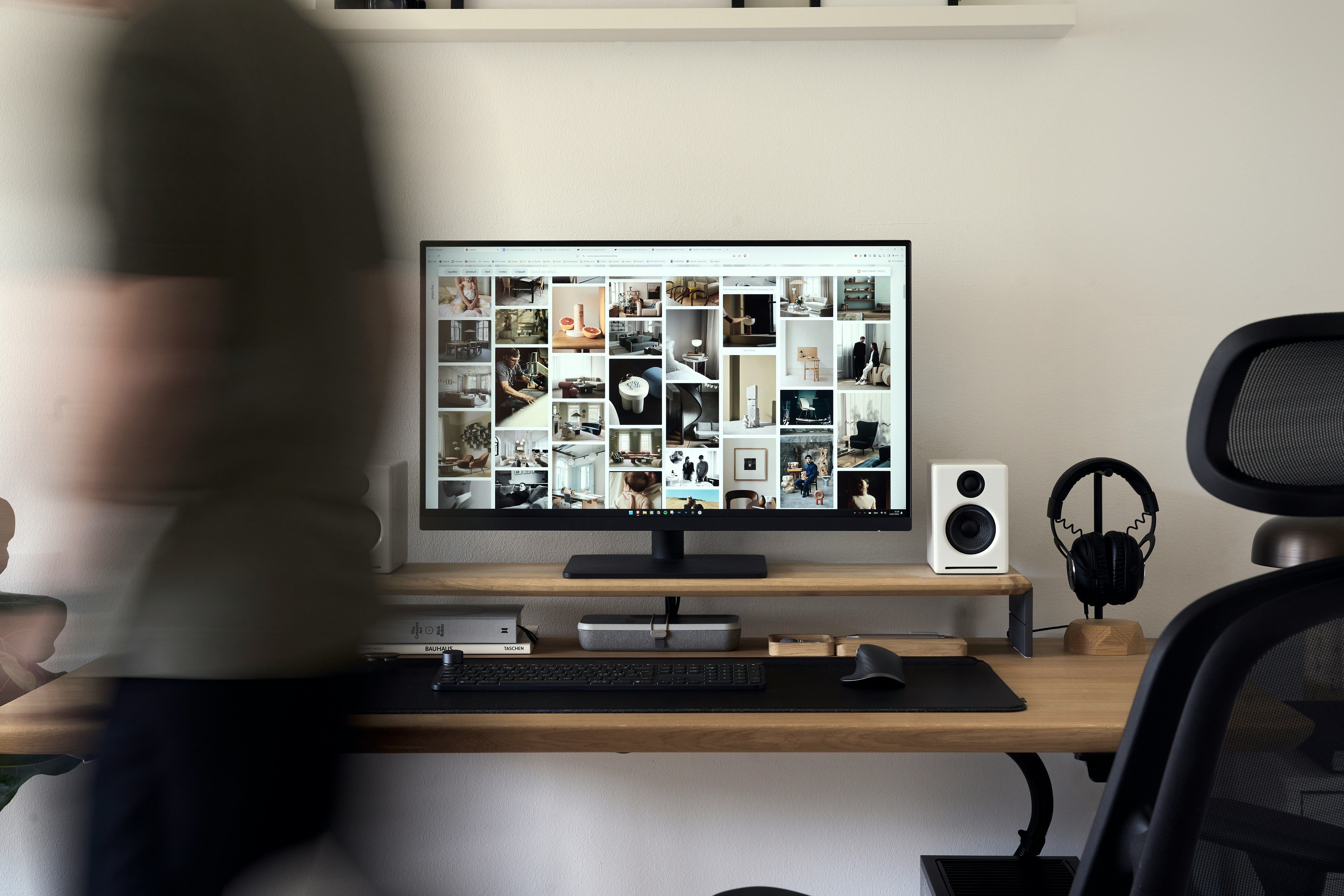 Desk with a monitor showing a grid of product images, speakers and headphones, a person blurred in motion beside it