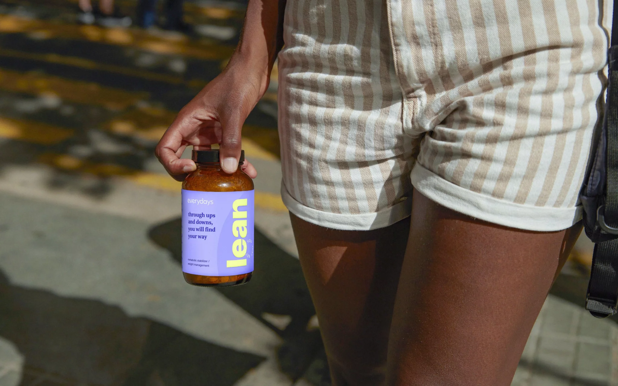 Person in striped shorts holding a purple-labelled 'lean' supplement jar on a sunny street, a DTC brand product