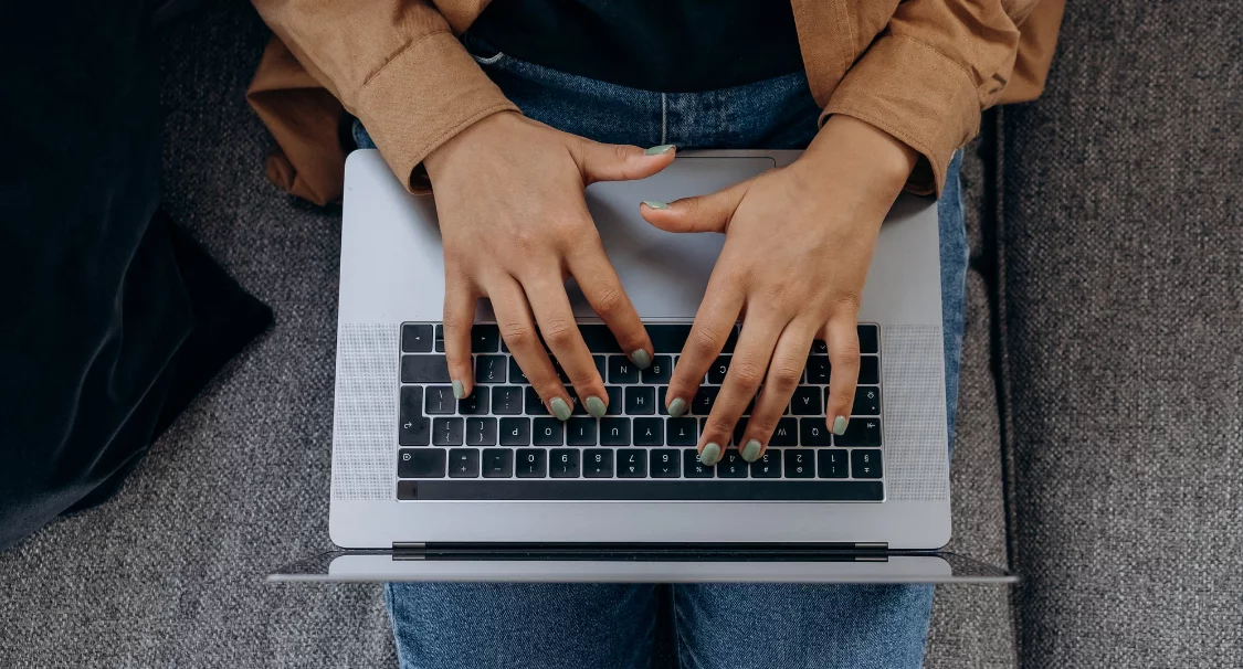 Top-down view of hands typing on a laptop resting on a person's lap on a grey sofa