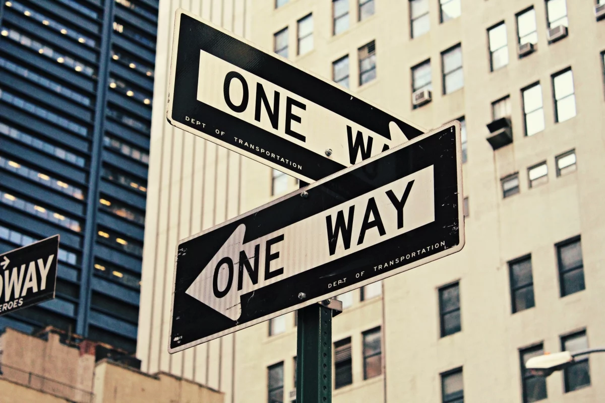 Two 'One Way' street signs pointing in opposite directions among city buildings, symbolising Shopify Plus vs. Magento