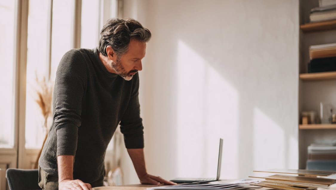 Man leaning over a desk with a laptop and paper reports in a sunlit office, pondering mismatched GA4 and Shopify revenue