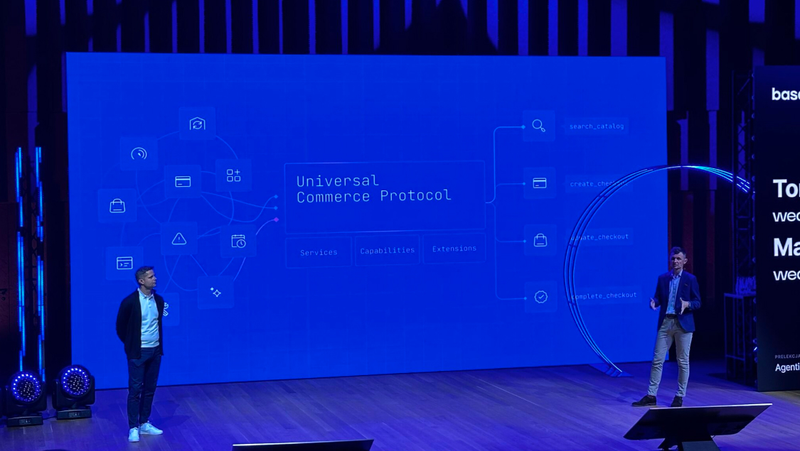 Two speakers on stage in front of a large blue screen presenting the 'Universal Commerce Protocol' diagram