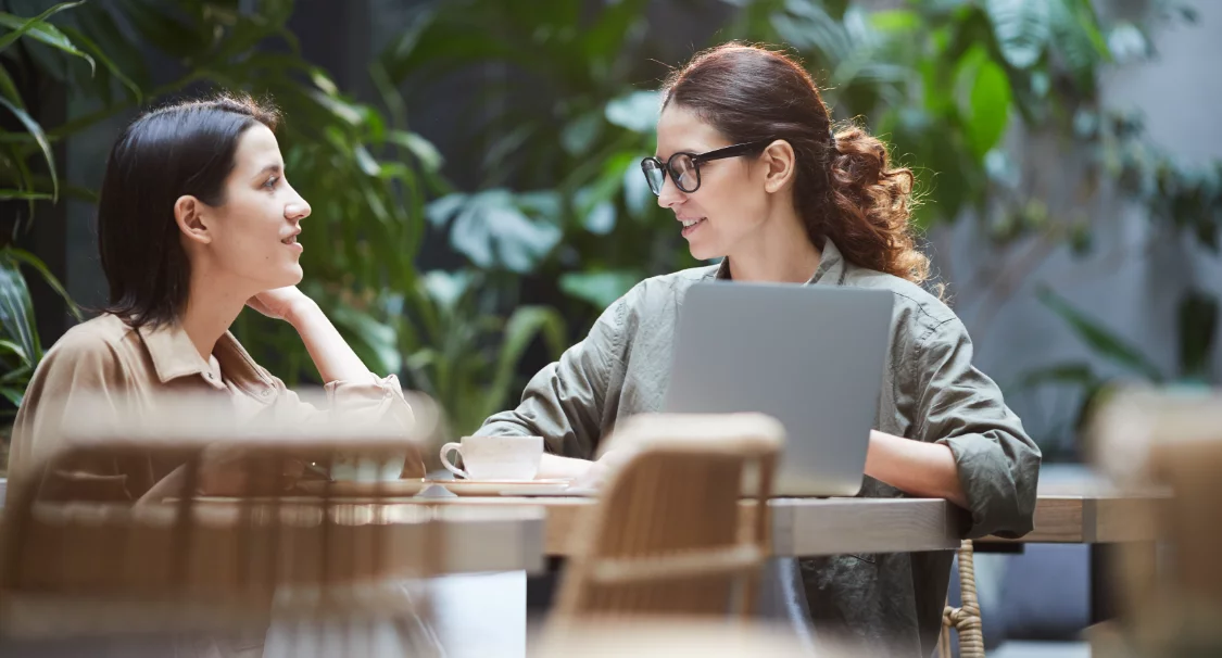 Two women talking at a café table with a laptop and coffee, discussing Shopify project pricing models