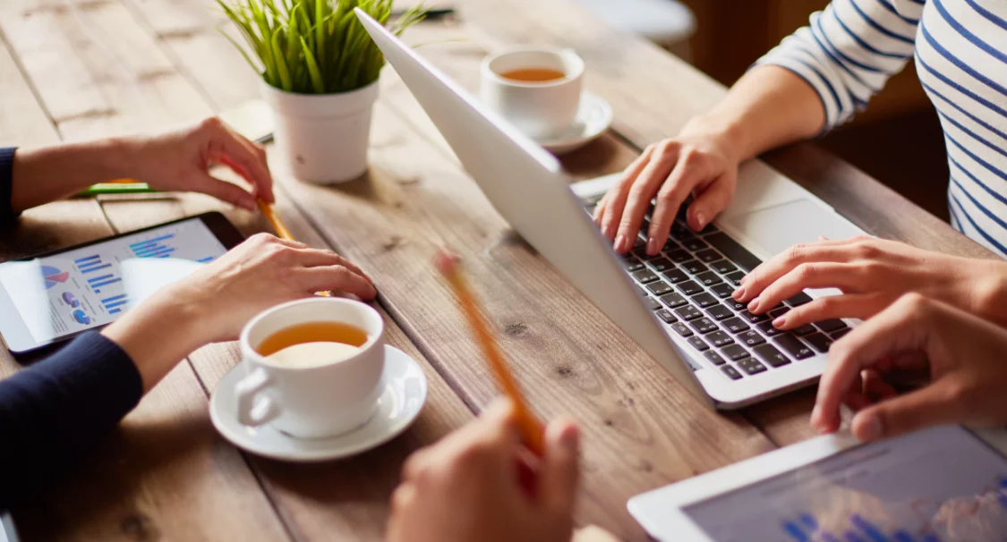 Hands working on a laptop and tablets with charts over a wooden table with tea cups, discussing e-commerce trends