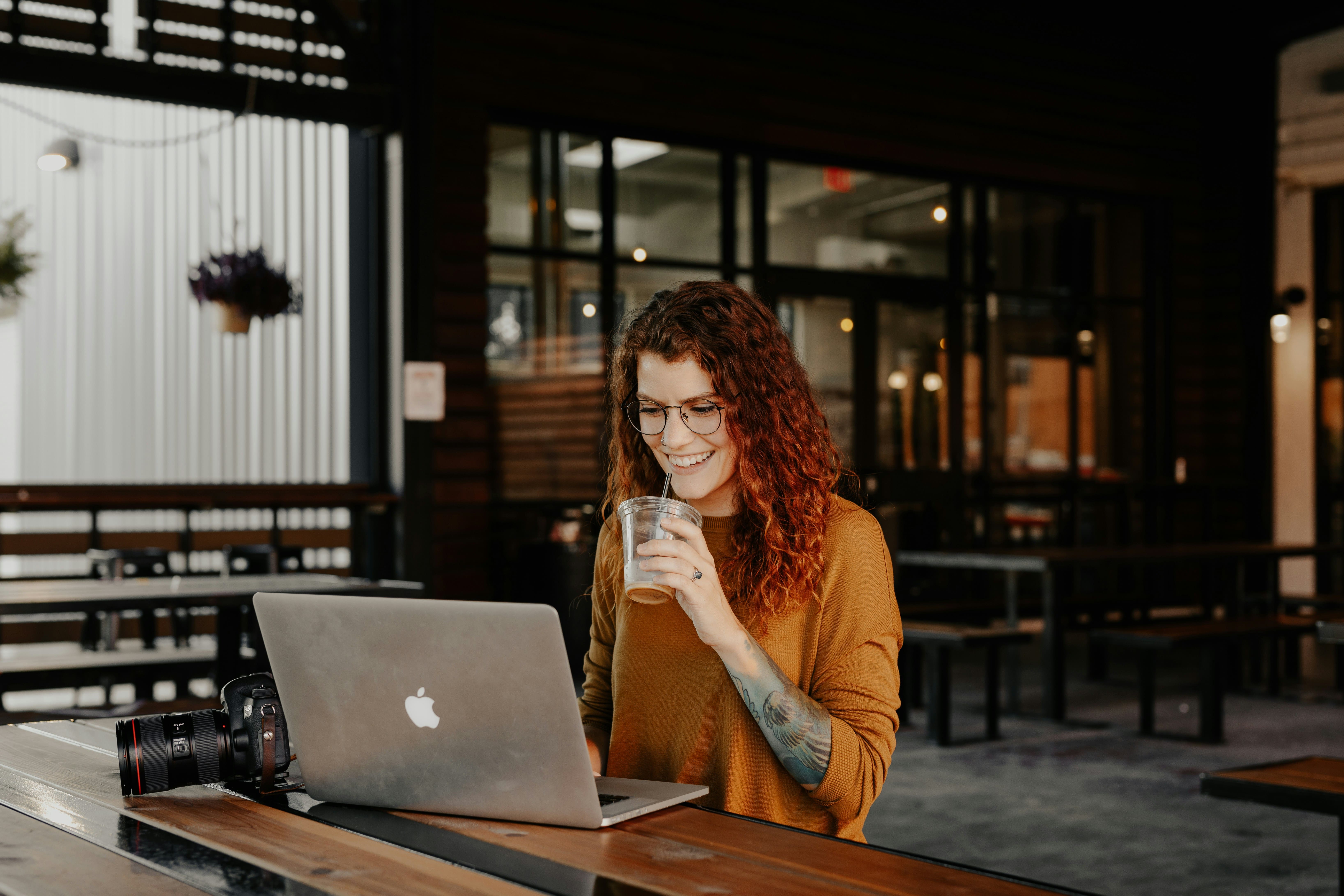 Smiling red-haired woman with a drink working on a laptop at a café table next to a camera, browsing product pages