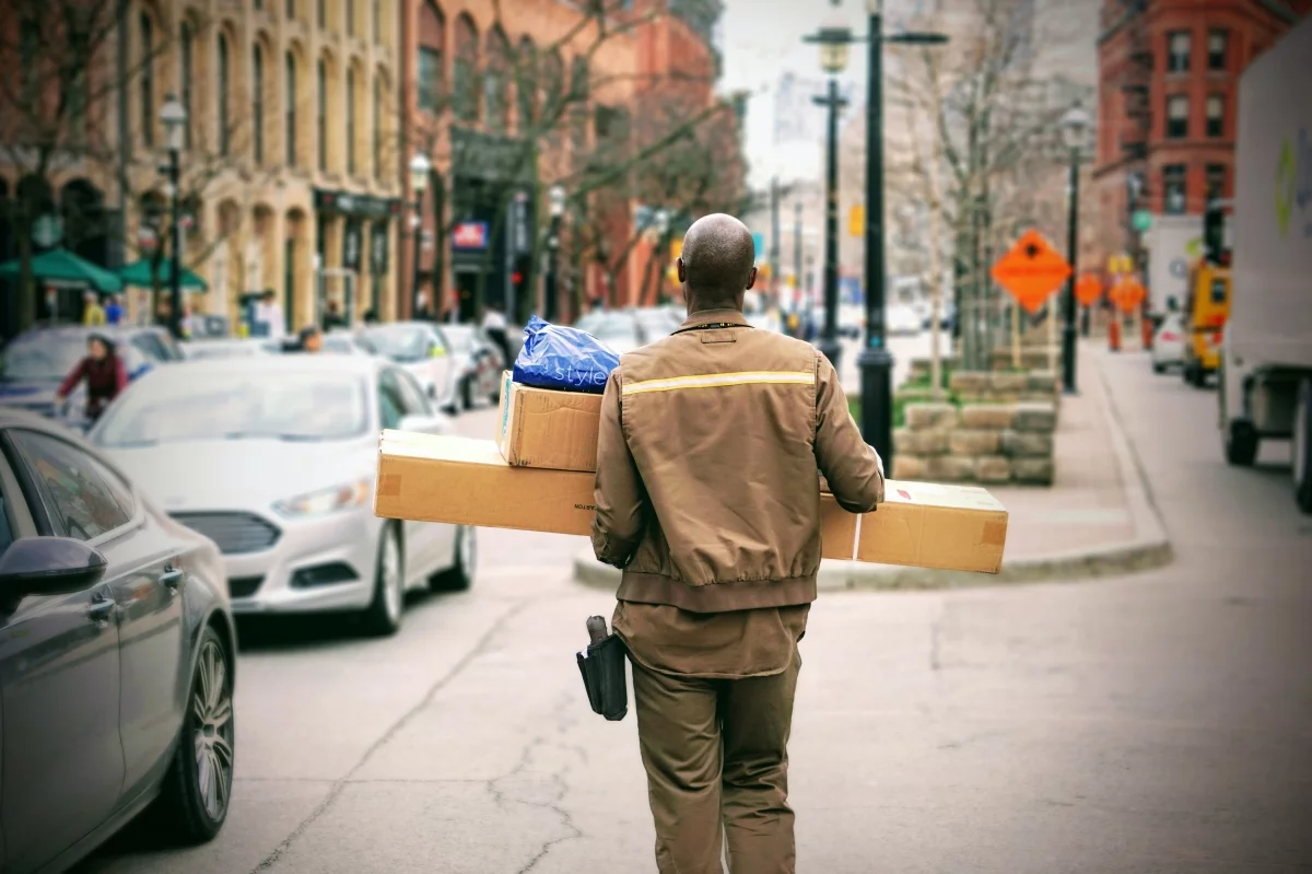 Courier in a brown uniform carrying cardboard boxes and a parcel along a street, illustrating delivery services